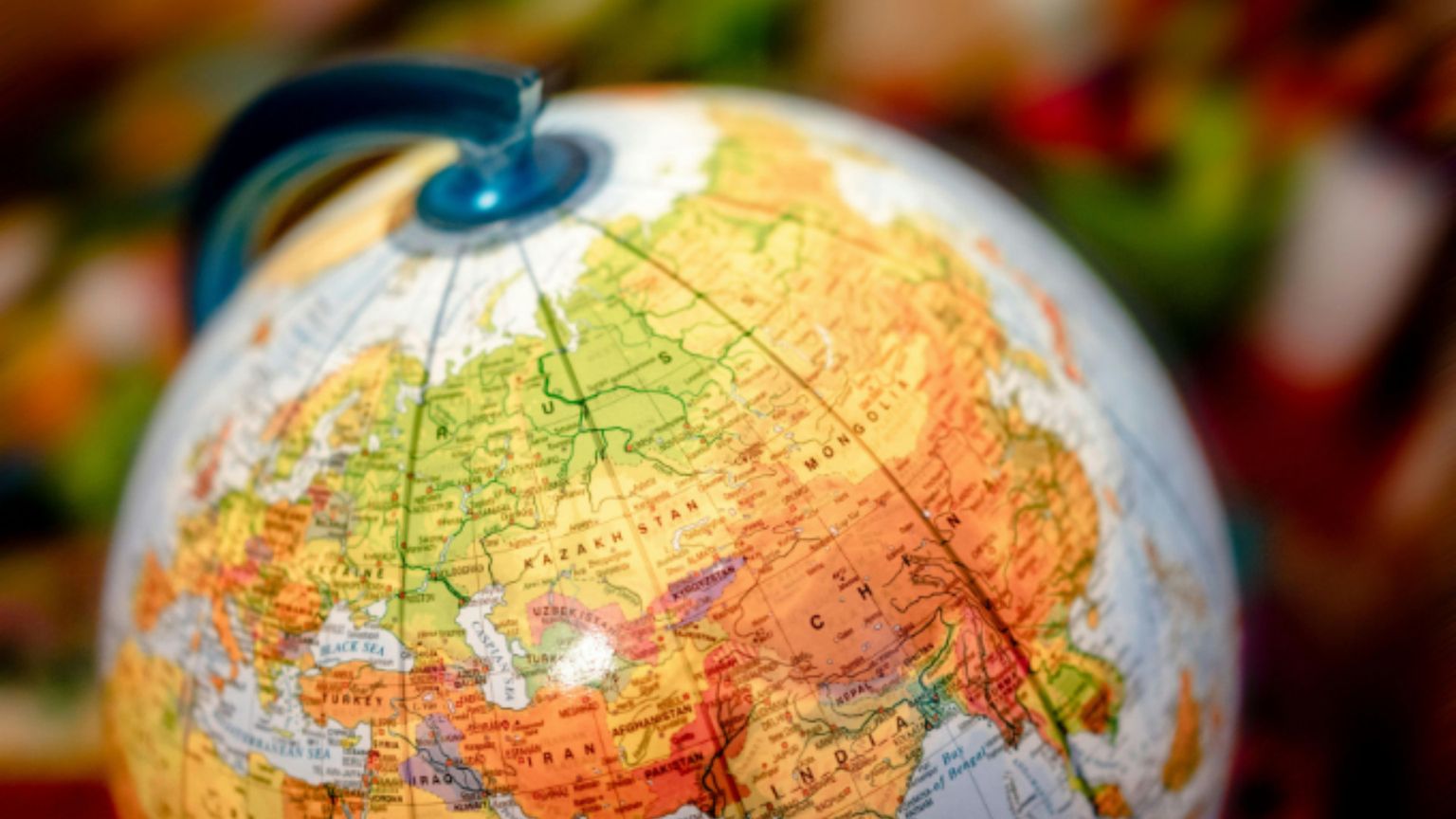 Un mappamondo colorato visto da vicino, leggermente inclinato, con la staffa blu in alto. A fuoco l’area tra Europa, Medio Oriente e Asia centrale; nomi e confini sono visibili. Lo sfondo è sfocato e multicolore