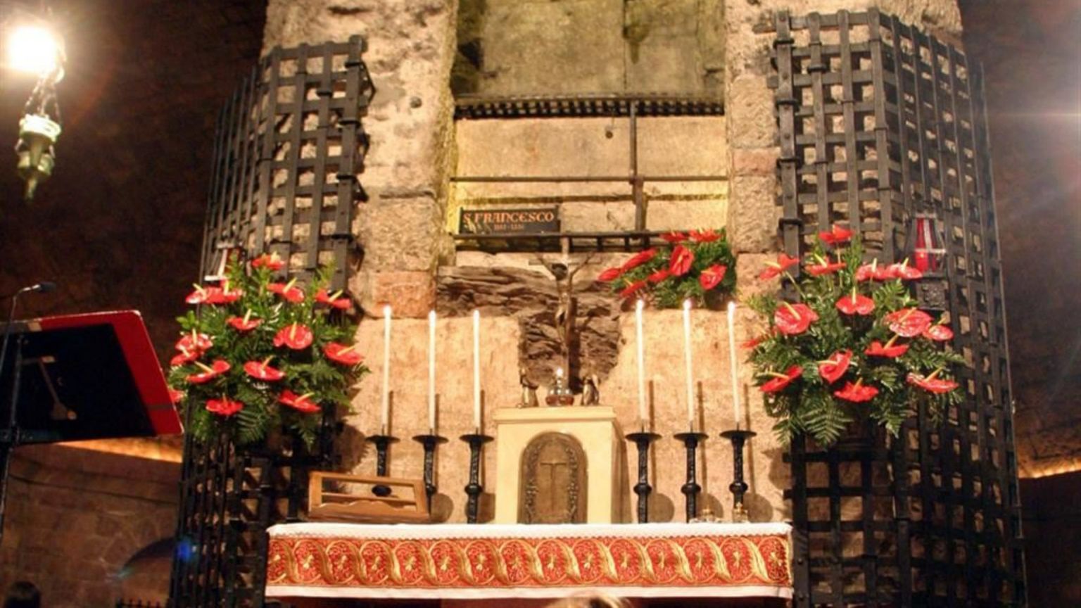 Altare in una cripta di pietra, illuminato in modo soffuso. Davanti ci sono sei candele bianche, un piccolo crocifisso e un rivestimento rosso ricamato. Ai lati, due grandi griglie metalliche con composizioni di fiori rossi. Al centro, in alto, una lapide