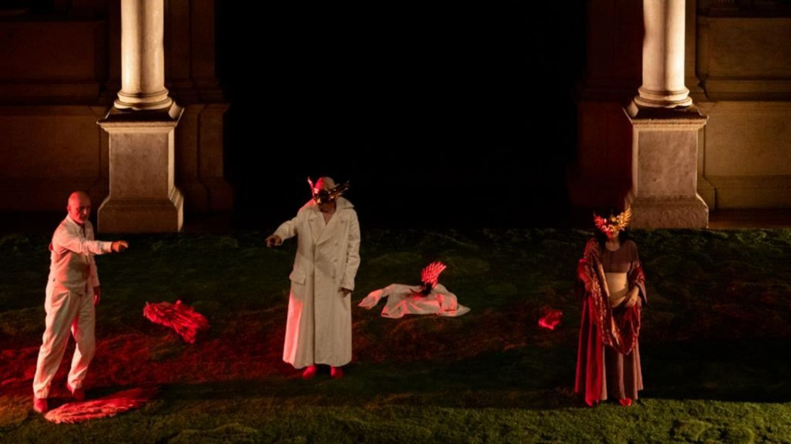 Scena teatrale in notturna: tre interpreti su un prato, incorniciati da due grandi colonne. Due indossano lunghi camici bianchi; due portano maschere dorate con piume