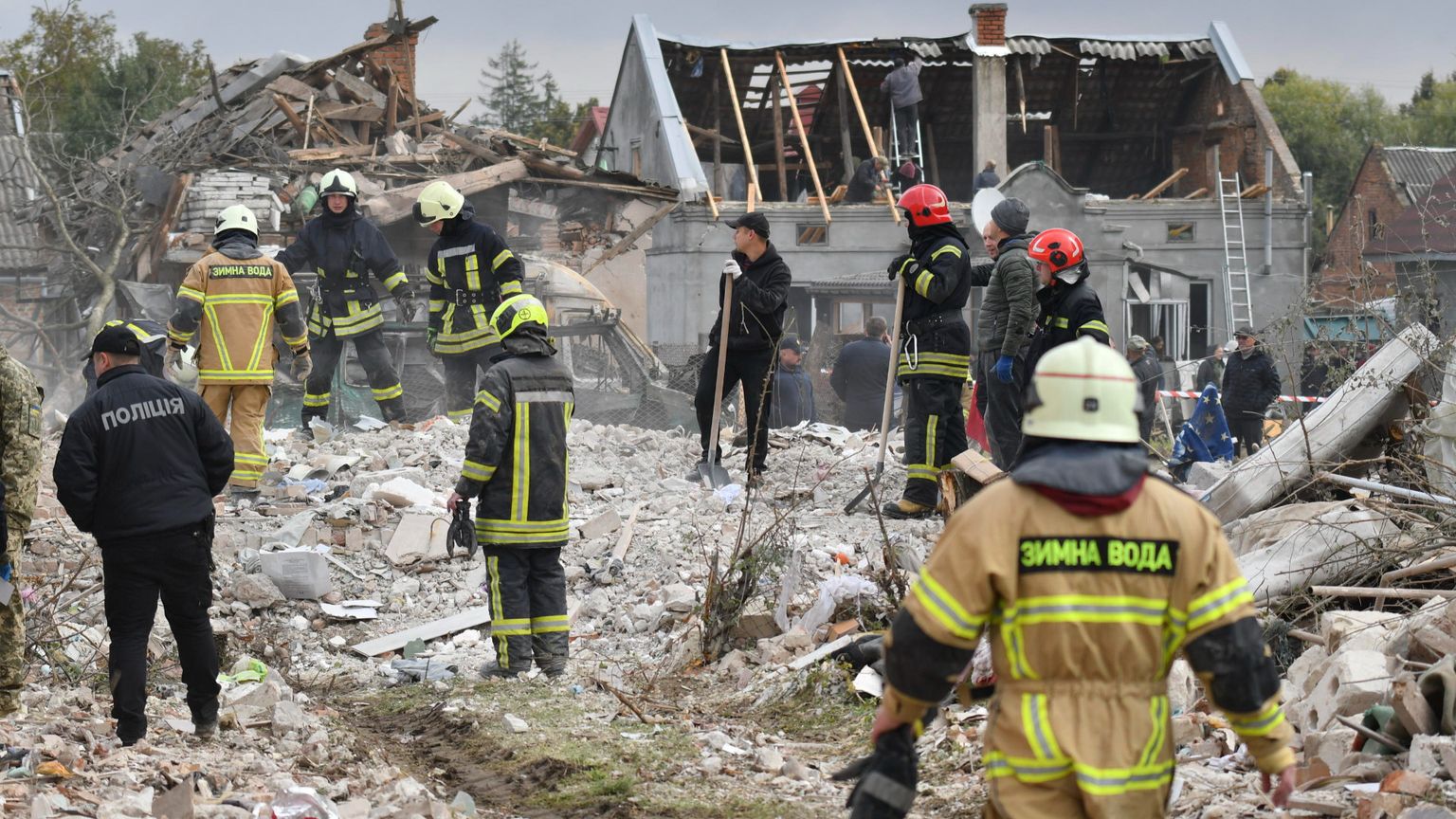 Uomini con tute da soccorritori, casco, guanti e pale in piedi su una distesa di detriti. Sullo sfondo un edificio con tetto a capanna scoperchiato
