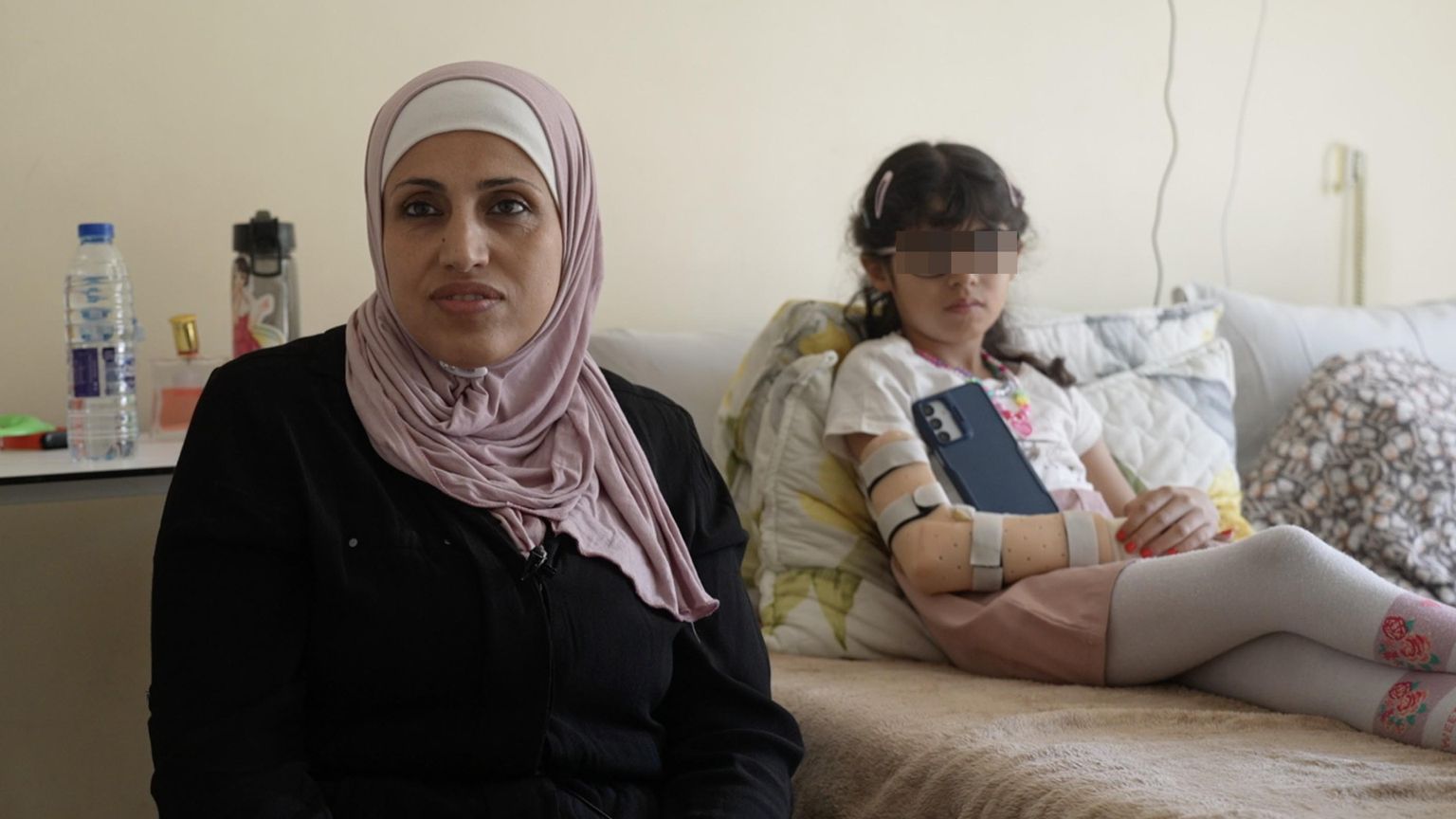 Una giovane donna vestita di nero, la testa coperta da un foulard color rosa antico, è seduta accanto a un lettino d'ospedale dov'è semisdraita, vestita, una bambina con gli occhiali, i capelli raccolti, lo smartphone. Un tutore sostiene il braccio destro