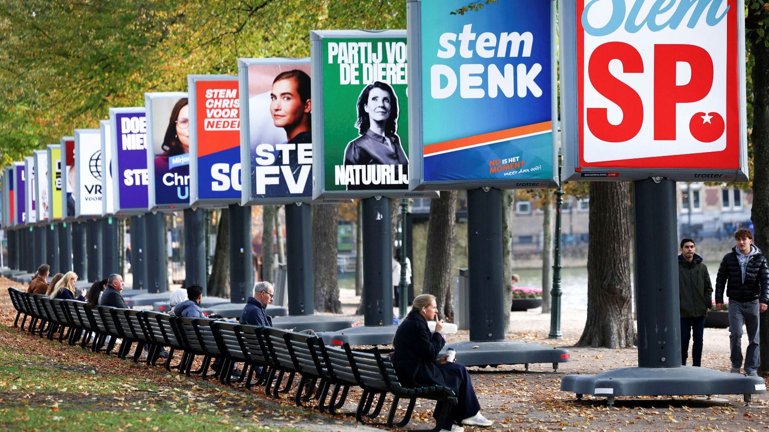 Una persona seduta su una panchina in un parco osserva dei grandi cartelloni elettorali con i simboli e gli slogan dei partiti