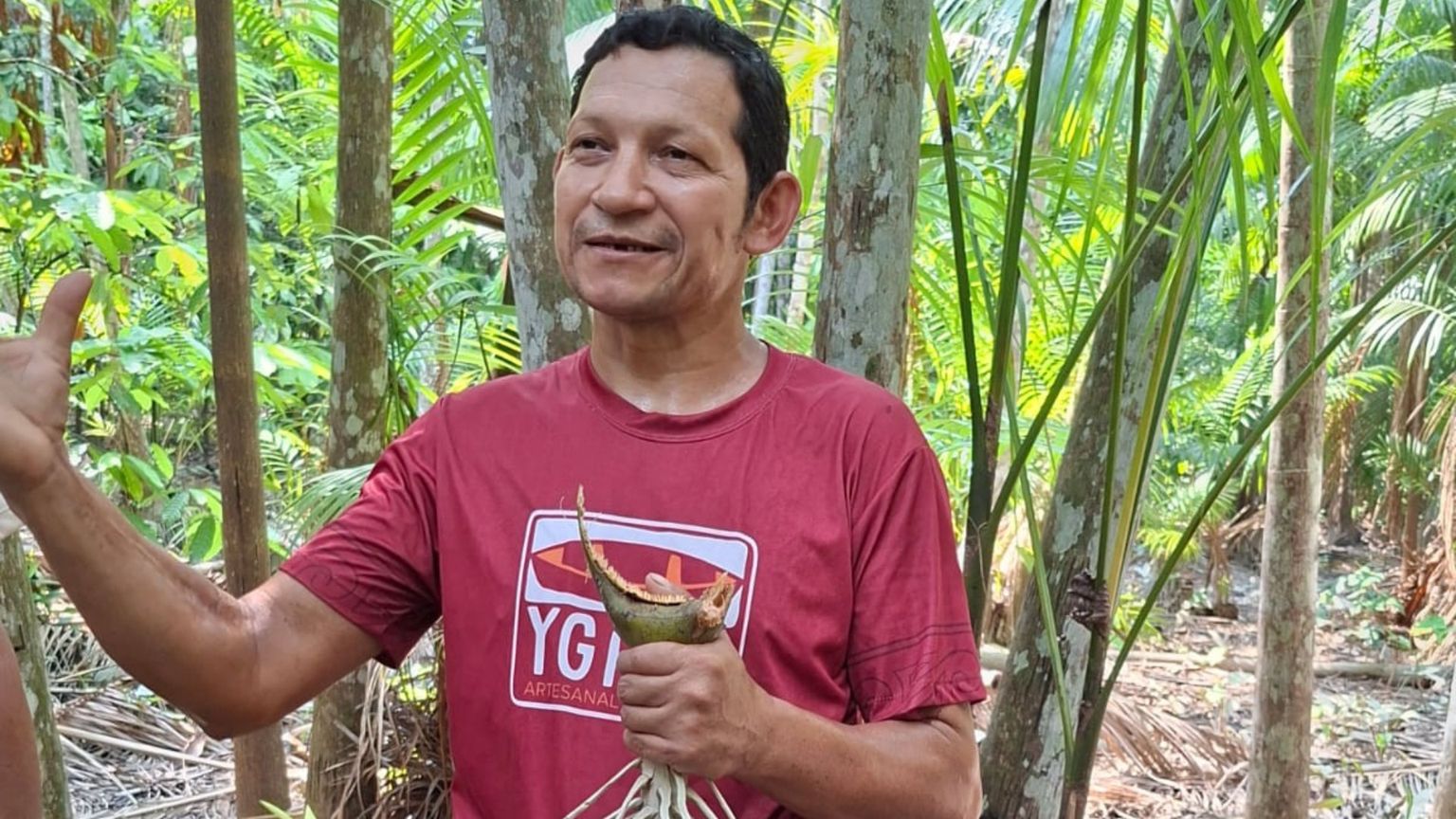 Un uomo di carnagione olivastra in maglietta rossa, in piedi e sorridente, in mezzo a una piantagione di giovani palme