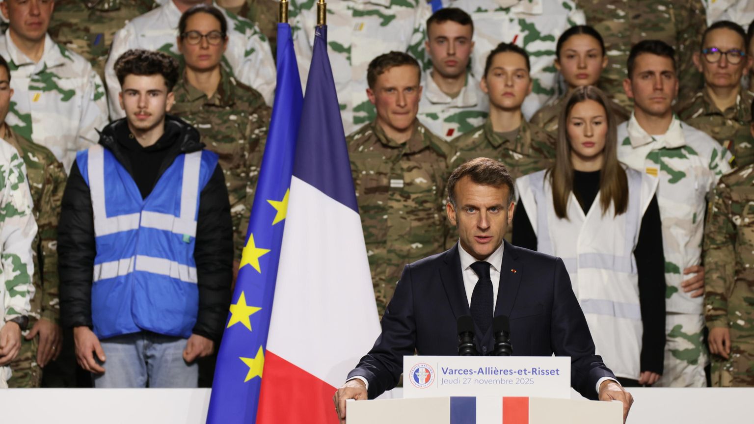 Il presidente francese Macron accanto alla bandiera francese e a quella dell'Unione Europea. Alle sue spalle uomini e donne in uniforme militare