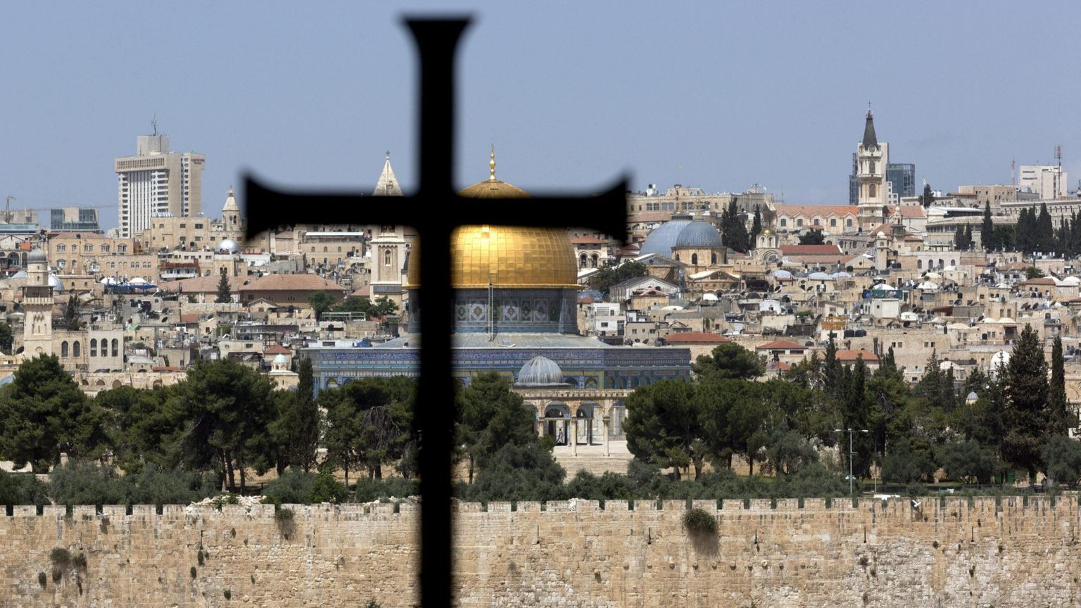 Una grande croce latina in primo piano, davanti al muro che cinge la Città Vecchia e oltre il quale spiccano l'oro della Cupola della Roccia, le cupole più basse del Santo Sepolcro e un campanile
