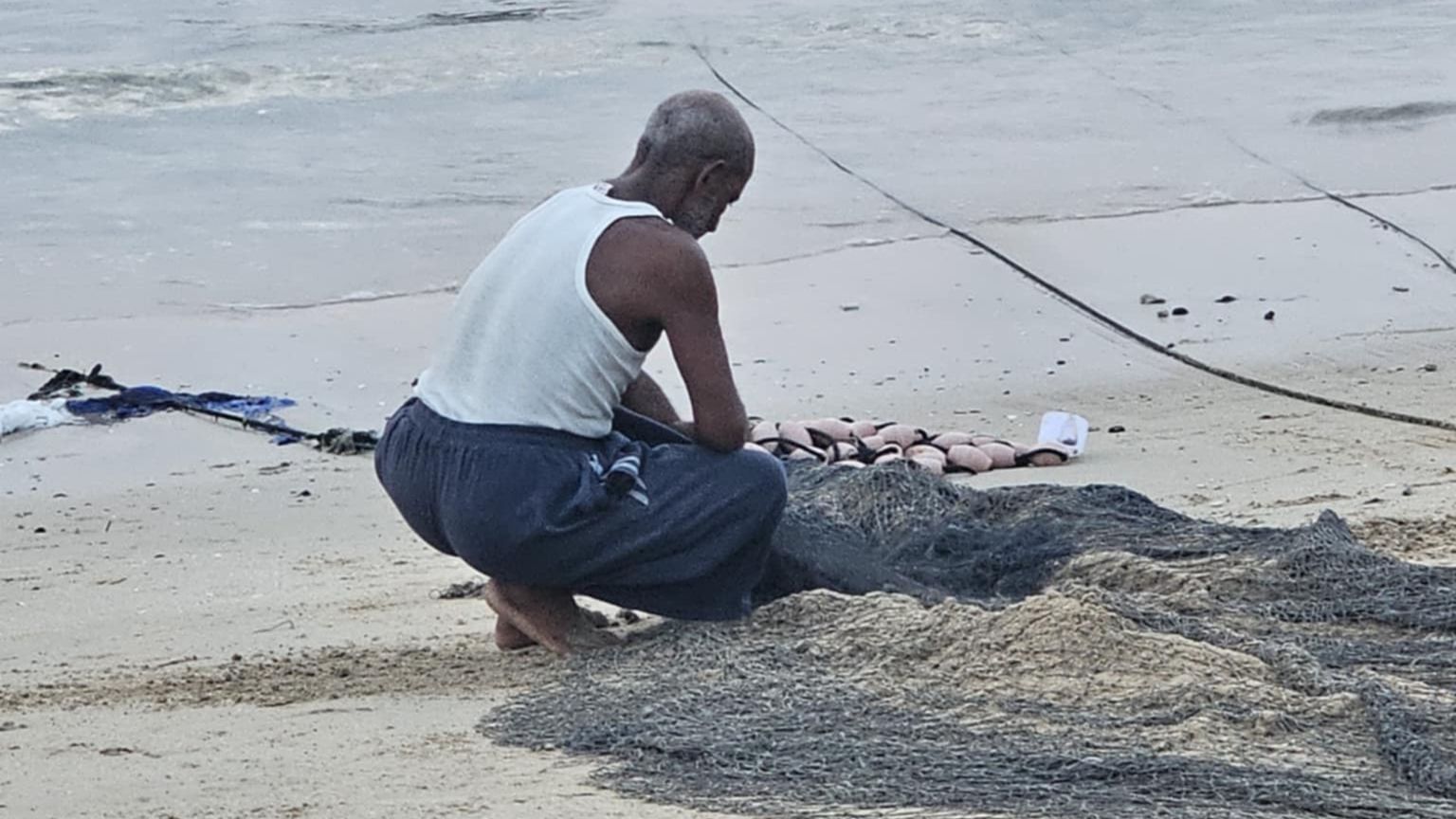 Un uomo in canottiera e a piedi nudi è accucciato sulla battigia davanti a reti da pesca
