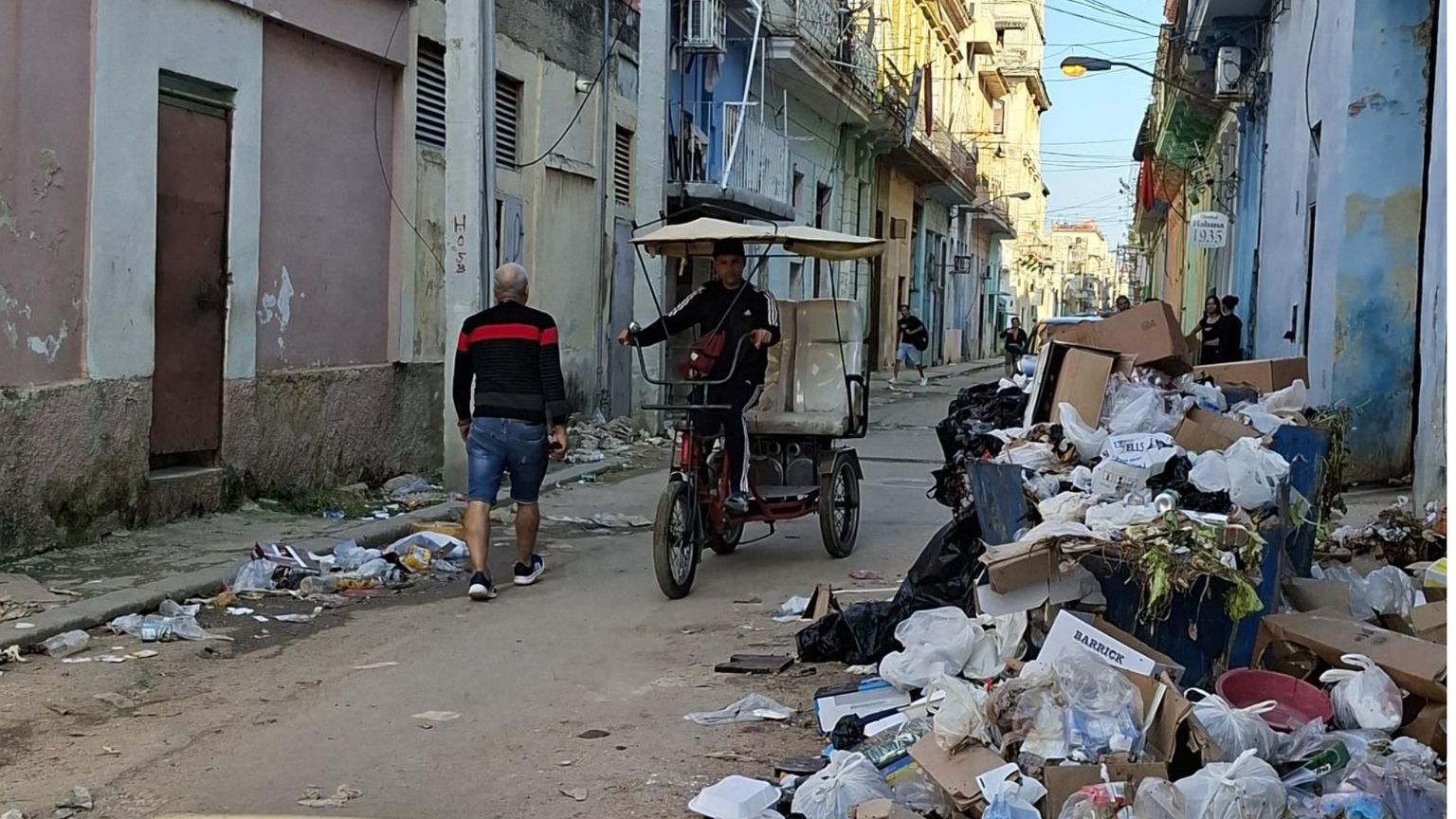 Nel centro dell'Avana semi-deserto un ragazzo guida un bici-taxi mentre un anziano si allontana. Una montagna di rifiuti giace sul ciglio della strada