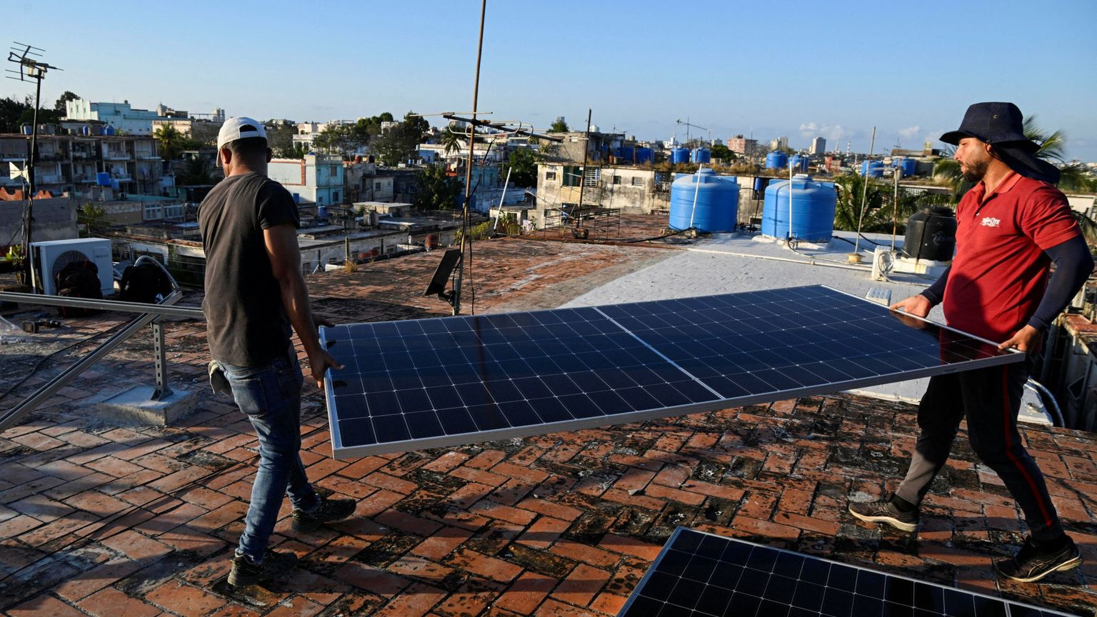 Due uomini sono sul tetto di una casa e trasportano un pannello solare. Dovranno installarlo in cima all'edificio. Sullo sfondo si intravedono altre case, alle cui sommità ci sono alcune cisterne per conservare l'acqua piovana.