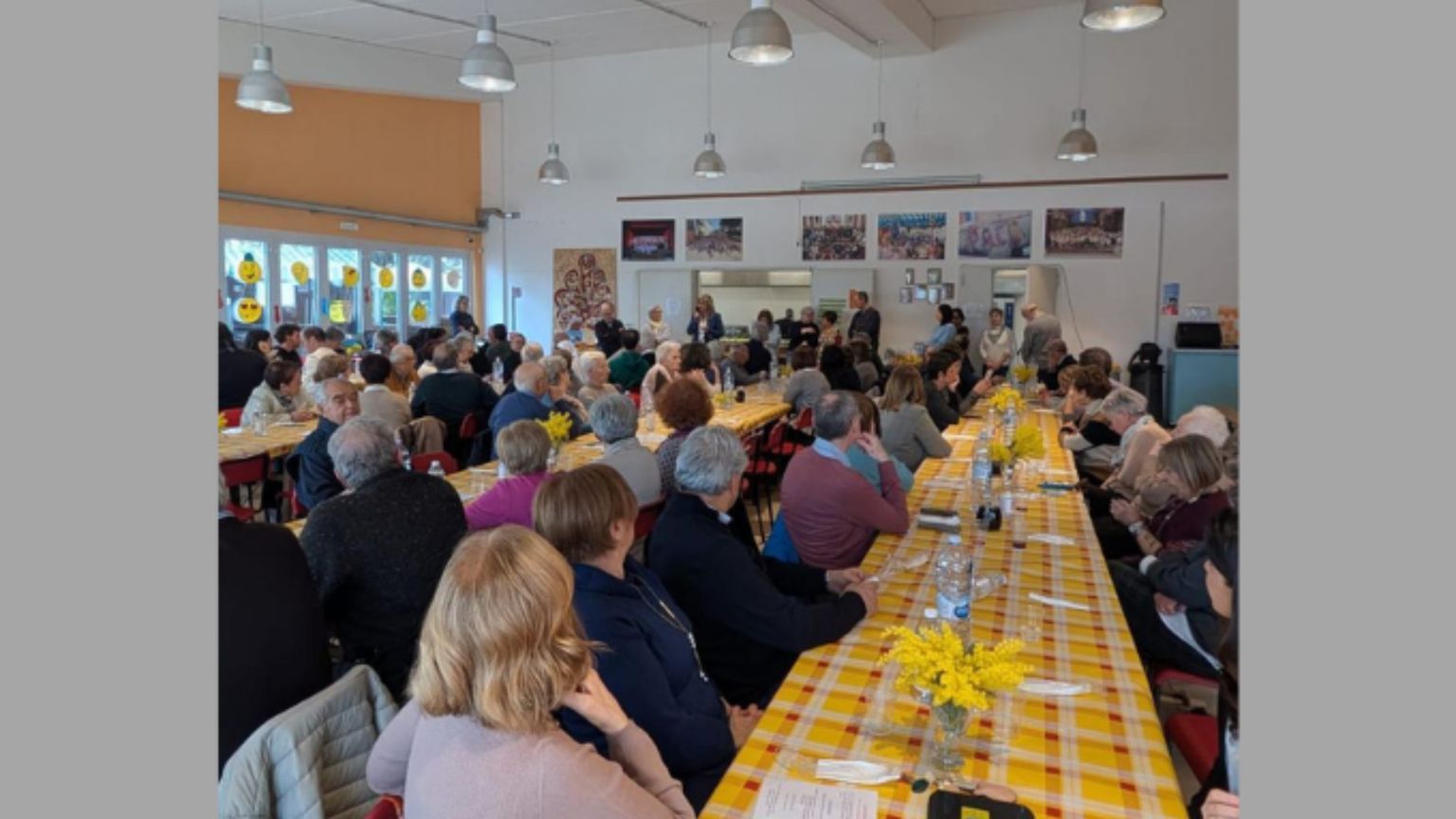 Più di cento persone sono sedute al tavolo; sono lunghe tavolate con tovaglie colorate. Si condivide il pasto.