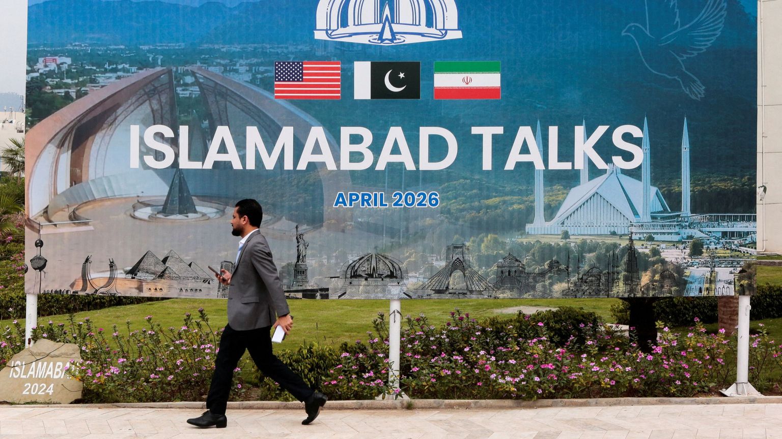 Un uomo passa davanti a un gigantesco cartellone con la scritta "Islamabad Talks" e le bandiere di Usa, Pakistan e Iran sormontate dall'emblema del Pakistan, una corona con mezzaluna