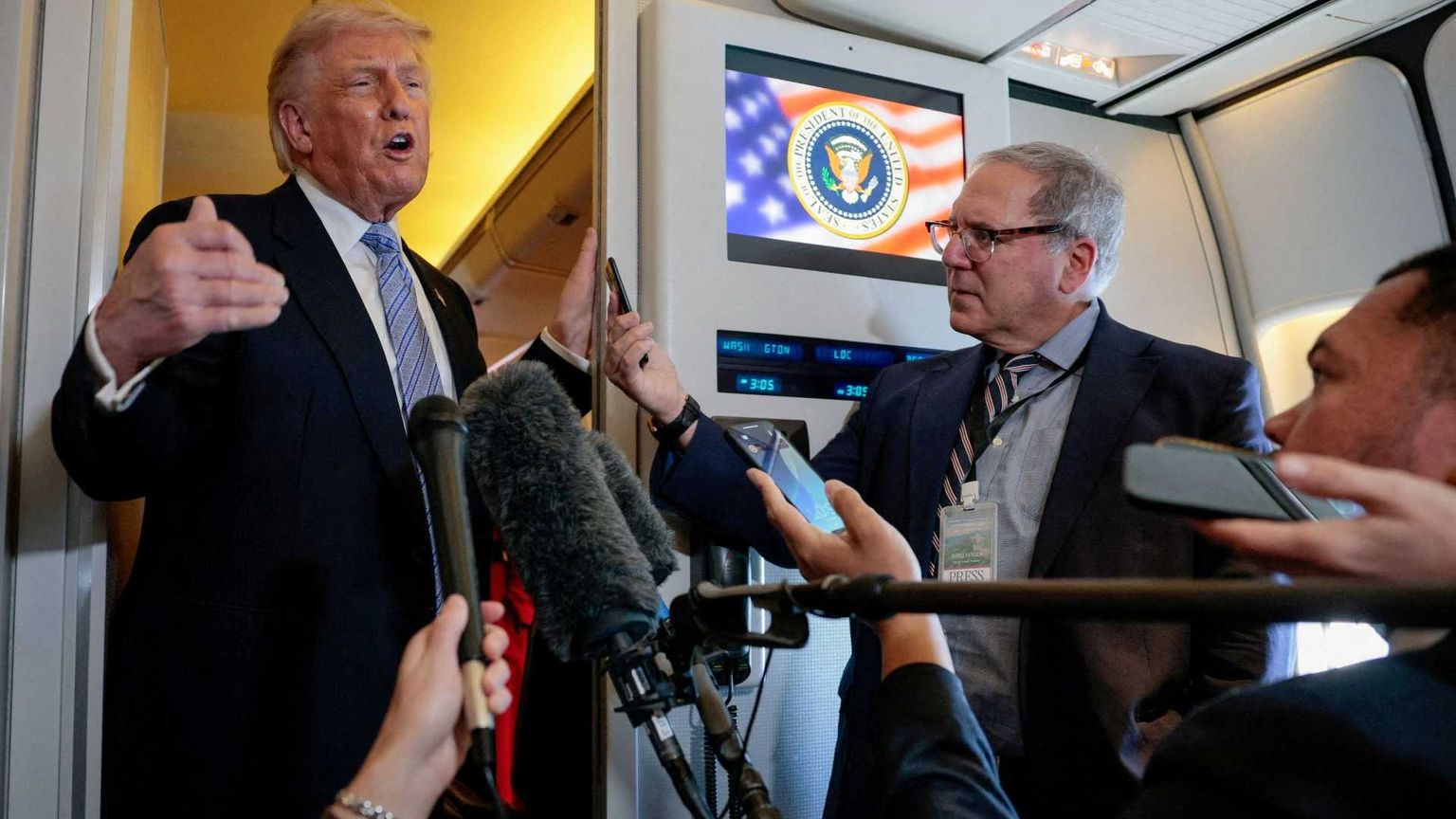 Trump in piedi davanti nel corridoio dell'aereo davanti a diversi microfoni tenuti dai giornalisti che lo accompagnano