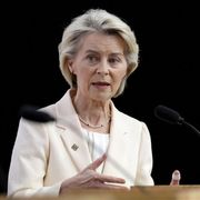 Una donna bionda, di mezza età, indossa un tailleur color crema e parla da un podio in legno. È ripresa a mezzobusto, con le mani sollevate mentre gesticola. Davanti a lei ci sono due microfoni neri; lo sfondo è completamente scuro, mettendo in risalto il