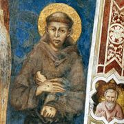 Un affresco medievale raffigura un monaco con tonsura e aureola dorata. Indossa un saio marrone e tiene le mani incrociate sul petto, davanti a uno sfondo blu intenso. A destra corre una cornice decorata in rosso e avorio, con un piccolo medaglione che mo