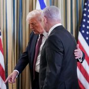 Due uomini in giacca e cravatta, di profilo, parlano da vicino in una stanza con tende a righe. Quello a sinistra, con capelli chiari, tiene una mano sulla spalla dell’altro e indica verso il pavimento; ai lati si vedono due bandiere degli Stati Uniti e,