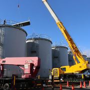 Il lungo braccio d'acciaio di una gru gialla si protende davanti alla prima di una serie di enormi cisterne cilindriche grigie, sotto il cielo azzurro