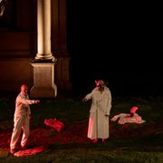 Scena teatrale in notturna: tre interpreti su un prato, incorniciati da due grandi colonne. Due indossano lunghi camici bianchi; due portano maschere dorate con piume