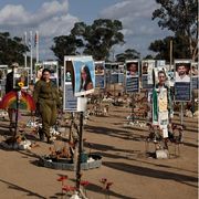 Campo commemorativo all’aperto: su terriccio chiaro sono disposti in file molti pannelli con foto e brevi testi delle vittime, circondati da piccoli memoriali con fiori, candele e oggetti personali