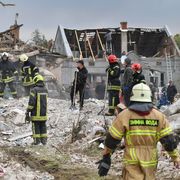 Uomini con tute da soccorritori, casco, guanti e pale in piedi su una distesa di detriti. Sullo sfondo un edificio con tetto a capanna scoperchiato