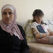 Una giovane donna vestita di nero, la testa coperta da un foulard color rosa antico, è seduta accanto a un lettino d'ospedale dov'è semisdraita, vestita, una bambina con gli occhiali, i capelli raccolti, lo smartphone. Un tutore sostiene il braccio destro