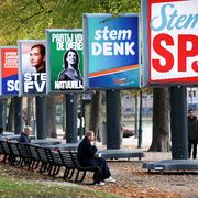 Una persona seduta su una panchina in un parco osserva dei grandi cartelloni elettorali con i simboli e gli slogan dei partiti