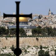 Una grande croce latina in primo piano, davanti al muro che cinge la Città Vecchia e oltre il quale spiccano l'oro della Cupola della Roccia, le cupole più basse del Santo Sepolcro e un campanile