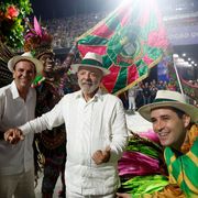 Lula vestito di bianco, con un panama in testa, in mezzo a due uomini col panama: quello a destra avvolto nei colori giallo e verde del Brasile. Sullo sfondo, ballerine in maschera e una bandiera