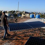 Due uomini sono sul tetto di una casa e trasportano un pannello solare. Dovranno installarlo in cima all'edificio. Sullo sfondo si intravedono altre case, alle cui sommità ci sono alcune cisterne per conservare l'acqua piovana.