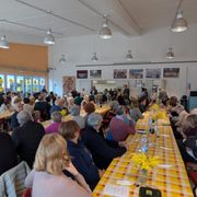 Più di cento persone sono sedute al tavolo; sono lunghe tavolate con tovaglie colorate. Si condivide il pasto.