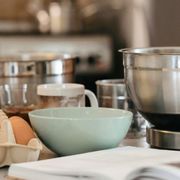 Preparazione di un nuovo piatto. Si vedono, su un tavolo, quattro contenitori, uno di ceramica e tre di acciaio, e diverse uova. In primo piano, sfocato, un libro di ricette