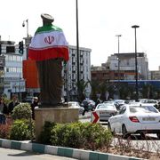 Una rotonda trafficata con, al centro, un'alta statua di un uomo con turbante: sulle spalle la bandiera dell'Iran