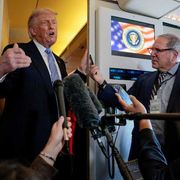 Trump in piedi davanti nel corridoio dell'aereo davanti a diversi microfoni tenuti dai giornalisti che lo accompagnano