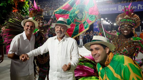 Lula vestito di bianco, con un panama in testa, in mezzo a due uomini col panama: quello a destra avvolto nei colori giallo e verde del Brasile. Sullo sfondo, ballerine in maschera e una bandiera