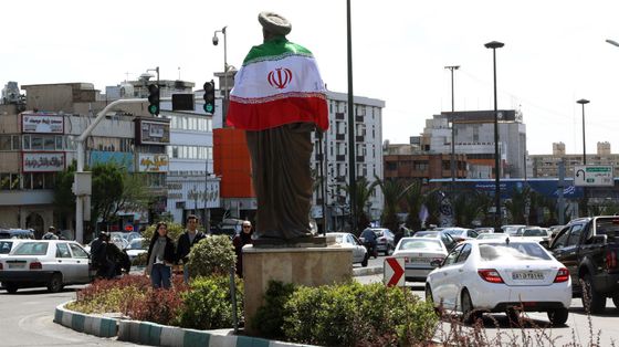 Una rotonda trafficata con, al centro, un'alta statua di un uomo con turbante: sulle spalle la bandiera dell'Iran