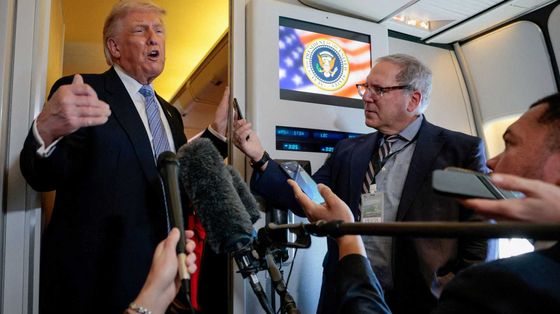 Trump in piedi davanti nel corridoio dell'aereo davanti a diversi microfoni tenuti dai giornalisti che lo accompagnano
