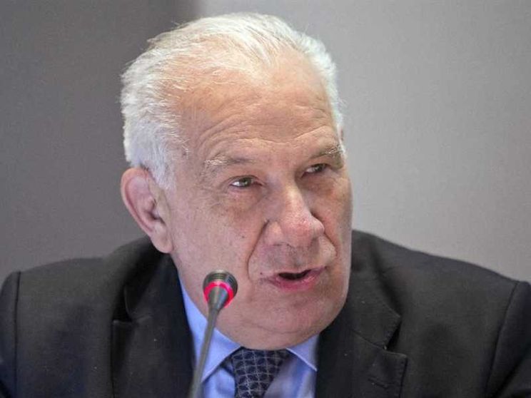 Primo piano di un uomo anziano dai capelli bianchi, in giacca e cravatta scura. È seduto a un tavolo e parla davanti a un microfono con una piccola luce rossa accesa. Lo sfondo è neutro; il suo volto è leggermente di profilo e l’espressione è concentrata.