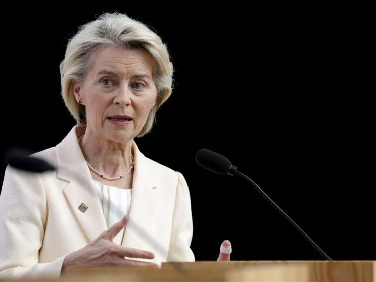 Una donna bionda, di mezza età, indossa un tailleur color crema e parla da un podio in legno. È ripresa a mezzobusto, con le mani sollevate mentre gesticola. Davanti a lei ci sono due microfoni neri; lo sfondo è completamente scuro, mettendo in risalto il