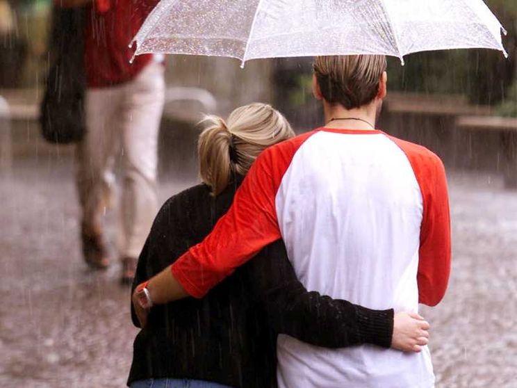 Due persone di spalle camminano abbracciate sotto un ombrello trasparente, sotto la pioggia. La strada è bagnata; sullo sfondo passano altri pedoni con ombrelli rossi. La persona a destra indossa una maglia bianca con maniche rosse, l’altra un maglione sc