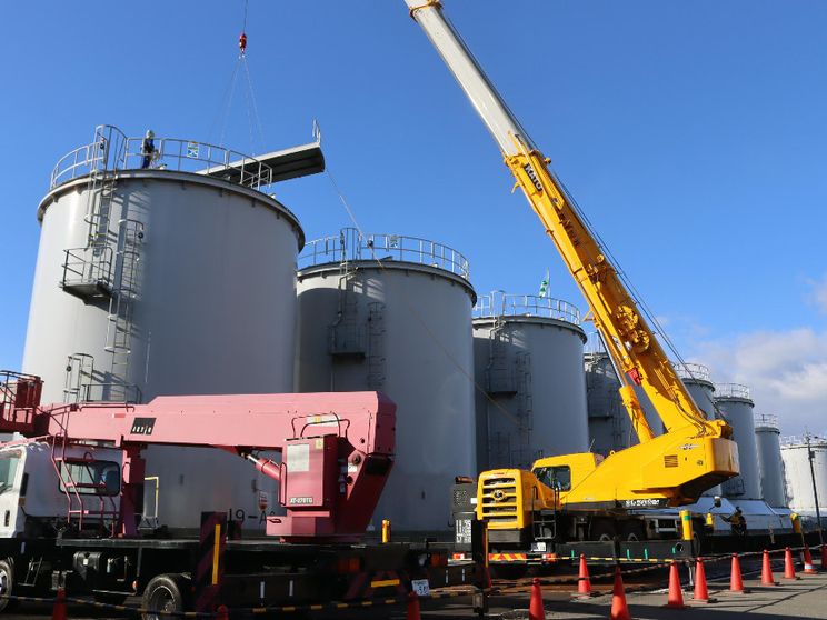 Il lungo braccio d'acciaio di una gru gialla si protende davanti alla prima di una serie di enormi cisterne cilindriche grigie, sotto il cielo azzurro