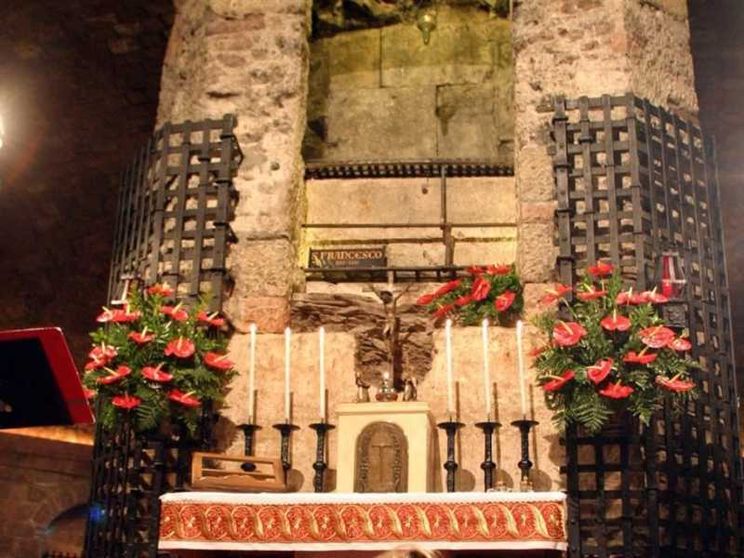 Altare in una cripta di pietra, illuminato in modo soffuso. Davanti ci sono sei candele bianche, un piccolo crocifisso e un rivestimento rosso ricamato. Ai lati, due grandi griglie metalliche con composizioni di fiori rossi. Al centro, in alto, una lapide