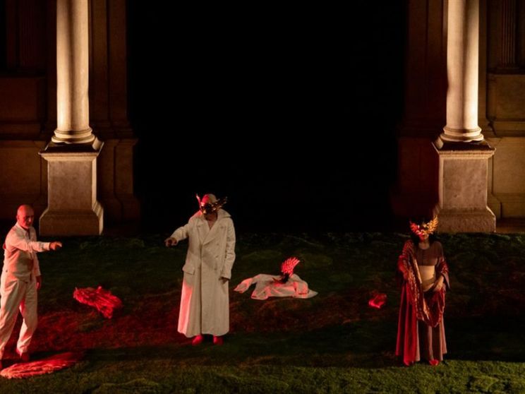 Scena teatrale in notturna: tre interpreti su un prato, incorniciati da due grandi colonne. Due indossano lunghi camici bianchi; due portano maschere dorate con piume