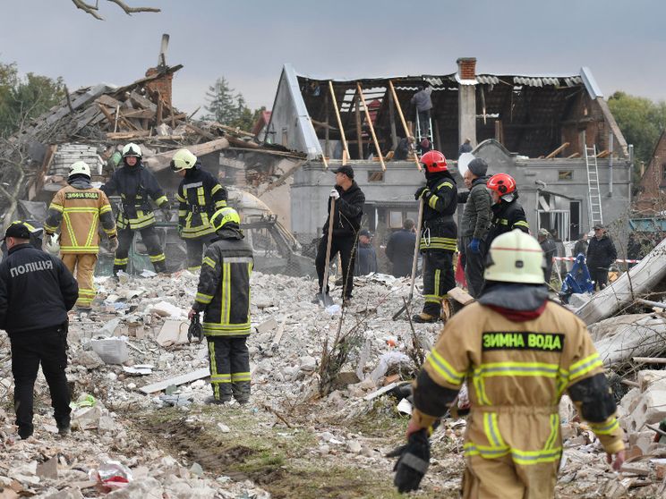 Uomini con tute da soccorritori, casco, guanti e pale in piedi su una distesa di detriti. Sullo sfondo un edificio con tetto a capanna scoperchiato
