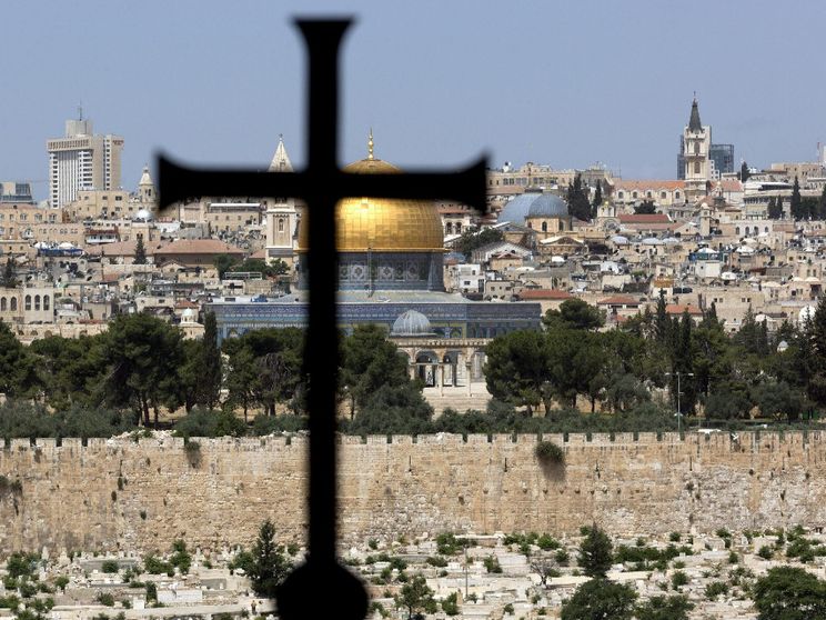 Una grande croce latina in primo piano, davanti al muro che cinge la Città Vecchia e oltre il quale spiccano l'oro della Cupola della Roccia, le cupole più basse del Santo Sepolcro e un campanile