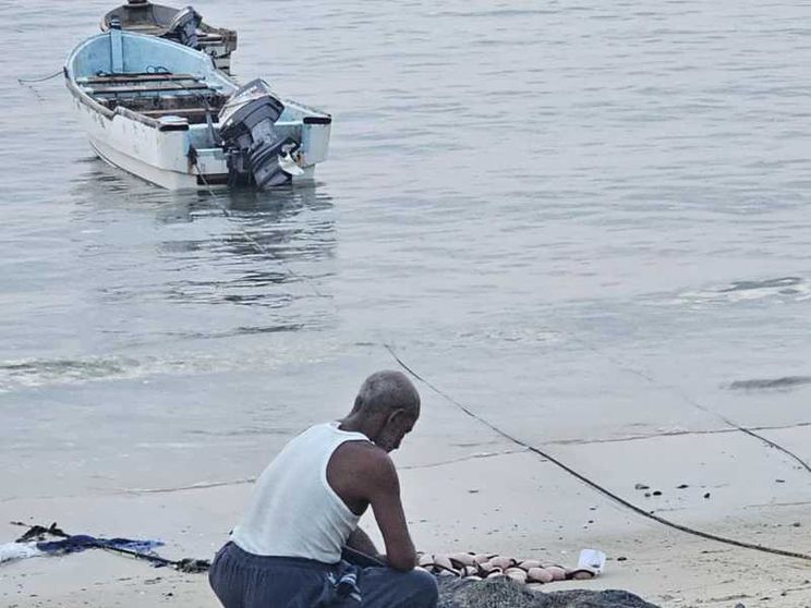 Un uomo in canottiera e a piedi nudi è accucciato sulla battigia davanti a reti da pesca