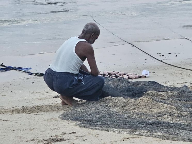 Un uomo in canottiera e a piedi nudi è accucciato sulla battigia davanti a reti da pesca