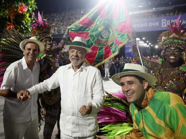 Lula vestito di bianco, con un panama in testa, in mezzo a due uomini col panama: quello a destra avvolto nei colori giallo e verde del Brasile. Sullo sfondo, ballerine in maschera e una bandiera