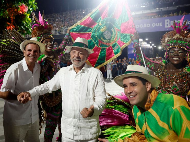 Lula vestito di bianco, con un panama in testa, in mezzo a due uomini col panama: quello a destra avvolto nei colori giallo e verde del Brasile. Sullo sfondo, ballerine in maschera e una bandiera
