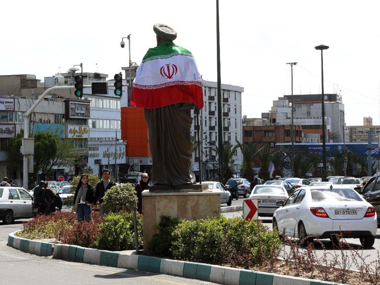 Una rotonda trafficata con, al centro, un'alta statua di un uomo con turbante: sulle spalle la bandiera dell'Iran