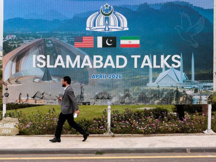 Un uomo passa davanti a un gigantesco cartellone con la scritta "Islamabad Talks" e le bandiere di Usa, Pakistan e Iran sormontate dall'emblema del Pakistan, una corona con mezzaluna