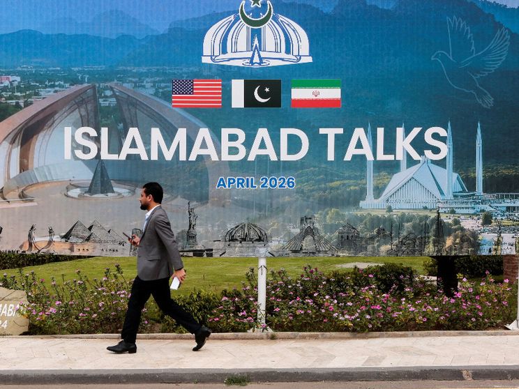 Un uomo passa davanti a un gigantesco cartellone con la scritta "Islamabad Talks" e le bandiere di Usa, Pakistan e Iran sormontate dall'emblema del Pakistan, una corona con mezzaluna