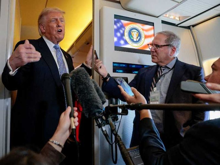 Trump in piedi davanti nel corridoio dell'aereo davanti a diversi microfoni tenuti dai giornalisti che lo accompagnano